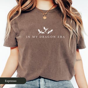 May include: Espresso-colored t-shirt with the phrase "IN MY DRAGON ERA" printed in white. Above the text are three white dragon silhouettes. The shirt has short sleeves and a crew neck.