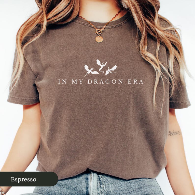 May include: Espresso-colored t-shirt with the phrase "IN MY DRAGON ERA" printed in white. Above the text are three white dragon silhouettes. The shirt has short sleeves and a crew neck.