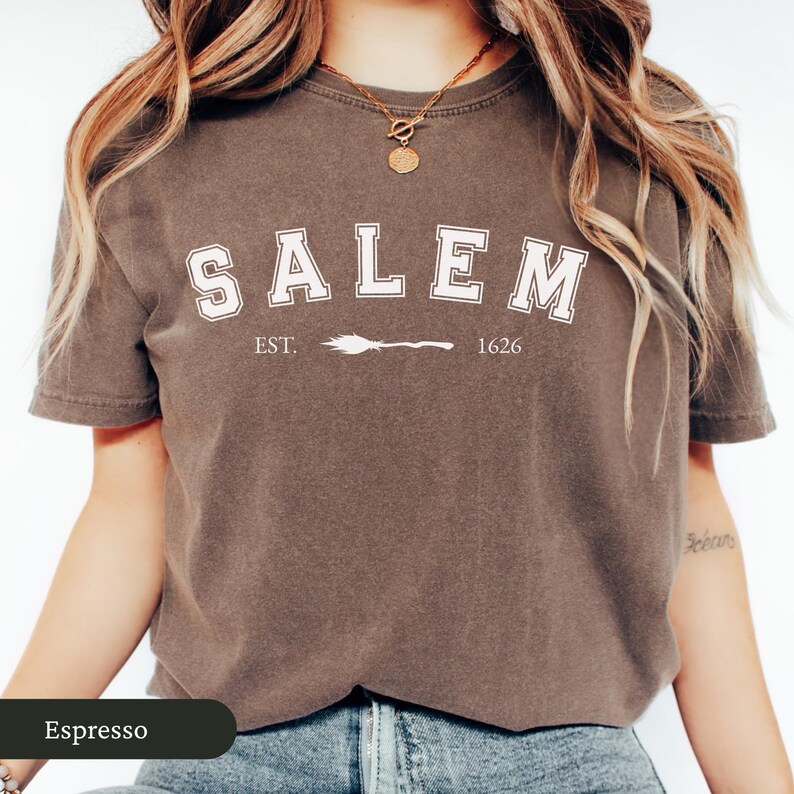 Peut inclure: T-shirt couleur expresso avec le mot "SALEM" en lettres majuscules blanches, soulign&eacute; d'une couleur plus fonc&eacute;e. En dessous du mot "SALEM" se trouve le texte "EST. 1626" avec un balai de sorci&egrave;re entre les deux. Le t-shirt est &agrave; col rond et &agrave; manches courtes.