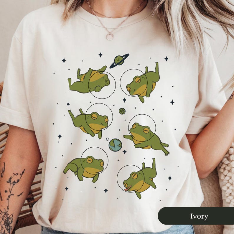 May include: Ivory-colored t-shirt featuring a whimsical design of green frogs in space suits floating among planets and stars. The frogs are in various poses, with a planet Earth and Saturn in the background. The shirt has short sleeves.
