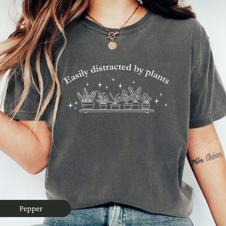 May include: Dark grey t-shirt with the text "Easily distracted by plants" in white. Below the text is a line drawing of potted plants on a shelf. The shirt is a solid colour and has short sleeves.