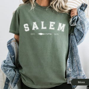 Peut inclure: T-shirt vert mousse avec le mot "SALEM" en blanc, en lettres arqu&eacute;es. En dessous, le texte "EST. 1626" avec un dessin de balai. Le t-shirt est &agrave; manches courtes et a un col rond.