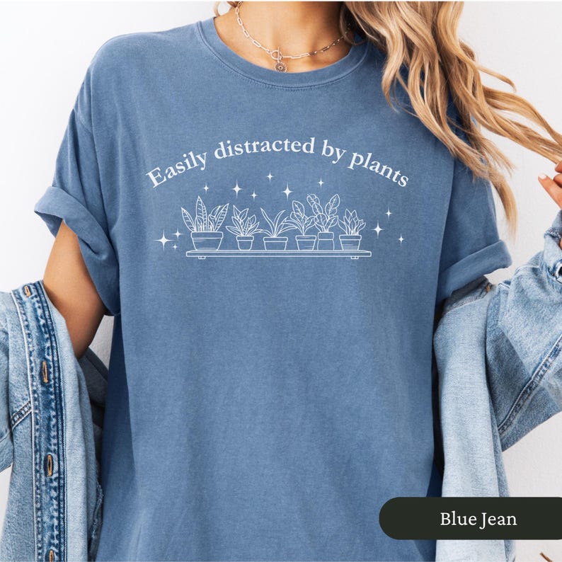 May include: A blue jean coloured t-shirt featuring the white text "Easily distracted by plants". Below the text is a shelf with potted plants and stars. The sleeves are rolled up.