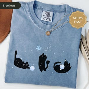 Puede incluir: Camiseta azul vaquero con un gr&aacute;fico juguet&oacute;n de tres gatos negros interactuando con una bola de nieve y un copo de nieve. La camiseta tiene cuello redondo y mangas cortas. La etiqueta dice "Comfort Colors". Un collar dorado con un colgante est&aacute; sobre la camiseta.