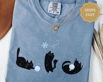 May include: Blue jean colored t-shirt with a playful graphic of three black cats interacting with a snowball and snowflake. The shirt has a crew neck and short sleeves. The tag reads "Comfort Colors". A gold necklace with a pendant is draped across the shirt.