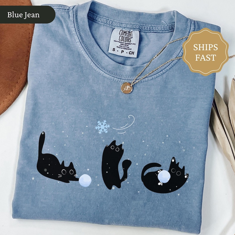 Puede incluir: Camiseta azul vaquero con un gr&aacute;fico juguet&oacute;n de tres gatos negros interactuando con una bola de nieve y un copo de nieve. La camiseta tiene cuello redondo y mangas cortas. La etiqueta dice "Comfort Colors". Un collar dorado con un colgante est&aacute; sobre la camiseta.