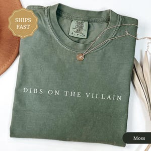 May include: A moss green t-shirt with the phrase "DIBS ON THE VILLAIN" printed in white. A gold necklace with a pendant rests on the shirt. The shirt is folded and has a tag that says "Comfort Colors".