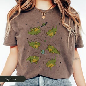 May include: Espresso-colored t-shirt featuring a whimsical design of green frogs in space suits, surrounded by planets and stars. The frogs are in various poses, creating a playful and unique graphic.