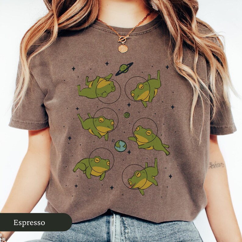 May include: Espresso-colored t-shirt featuring a whimsical design of green frogs in space suits, surrounded by planets and stars. The frogs are in various poses, creating a playful and unique graphic.