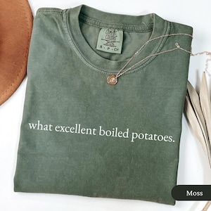 May include: A moss green t-shirt with the phrase "what excellent boiled potatoes." printed in white. A gold necklace with a pendant rests on the shirt. The shirt is folded and has a tag that reads "Comfort Colors".