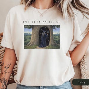 Op de afbeelding: Ivoor t-shirt met de tekst "I'LL BE IN MY OFFICE" boven een fantasie-illustratie. De illustratie toont een tovenaar in een donkerblauwe jurk in een boomdeur, met een gloeiende staf.
