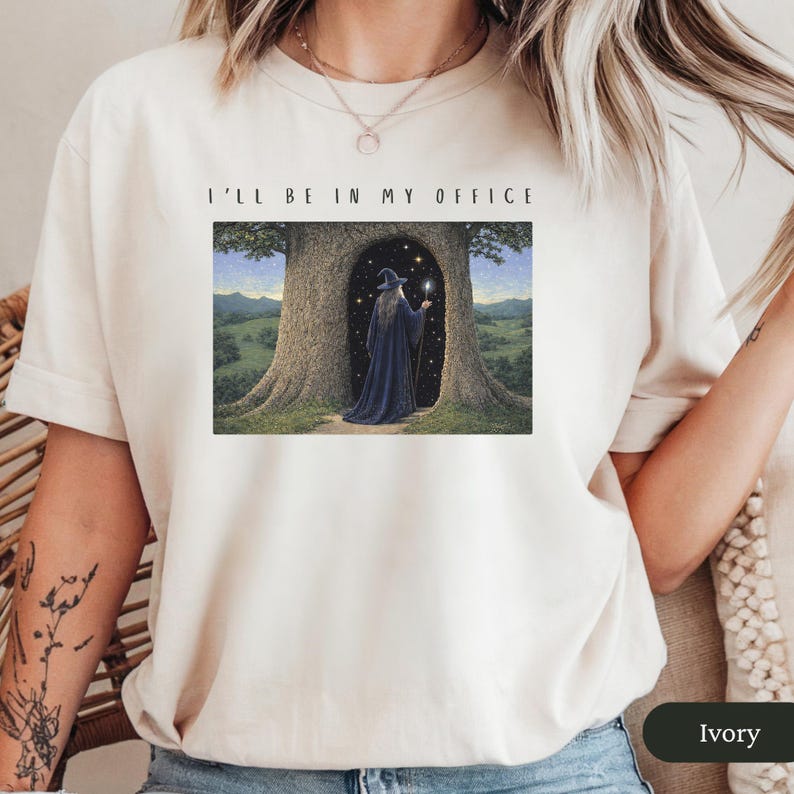 Op de afbeelding: Ivoor t-shirt met de tekst "I'LL BE IN MY OFFICE" boven een fantasie-illustratie. De illustratie toont een tovenaar in een donkerblauwe jurk in een boomdeur, met een gloeiende staf.