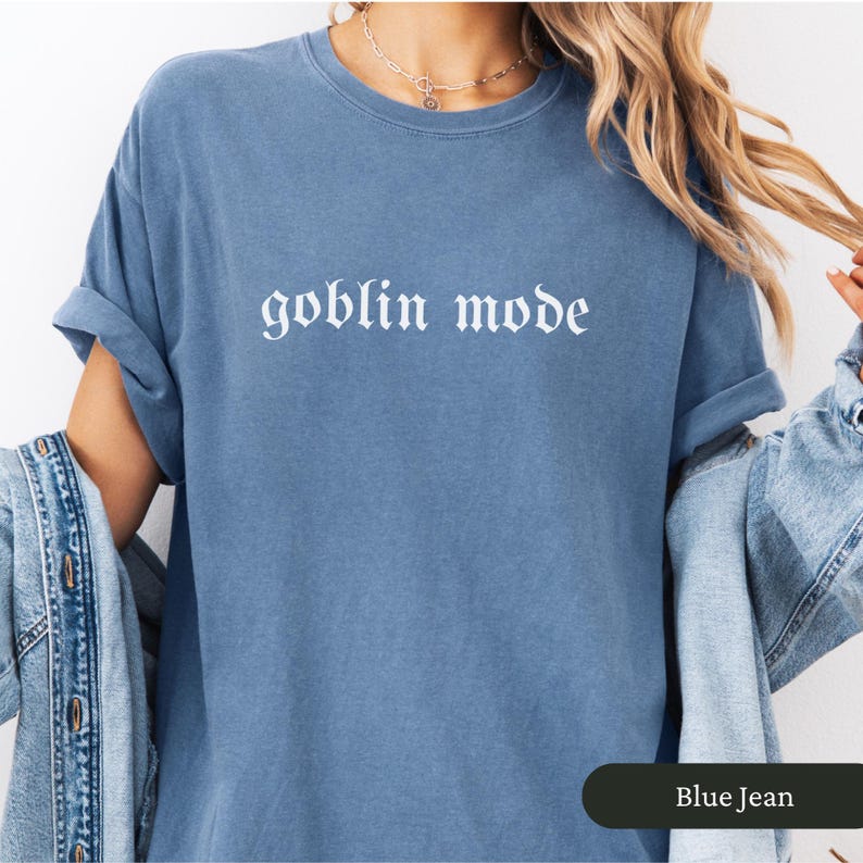 May include: Blue jean-colored t-shirt with the words "goblin mode" in white gothic font. The shirt has rolled-up sleeves and a relaxed fit. The text "Blue Jean" is in a dark green rectangle at the bottom.