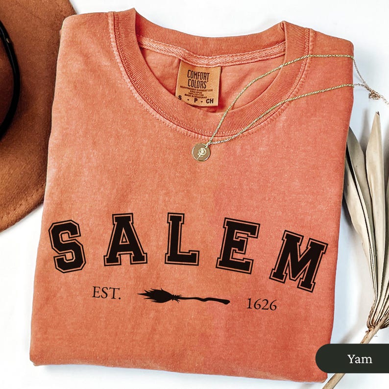 Peut inclure: Un sweat-shirt Comfort Colors de couleur corail avec le mot "SALEM" en lettres noires, un dessin de balai et le texte "EST. 1626." Un collier dor&eacute; avec un pendentif repose sur le t-shirt. Le t-shirt est de taille S.