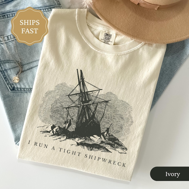 May include: Ivory-colored t-shirt with a black graphic of a ship in a storm, with the text "I RUN A TIGHT SHIPWRECK". The shirt is folded and placed on a light blue denim surface, with a necklace and a brown hat.
