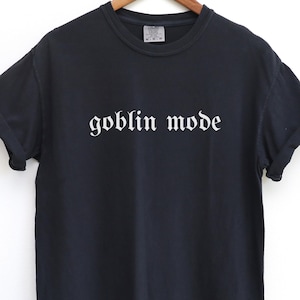May include: Black t-shirt with the words "goblin mode" printed in white gothic font across the front. The shirt has short sleeves and a crew neck. The fabric appears to be a soft cotton blend.