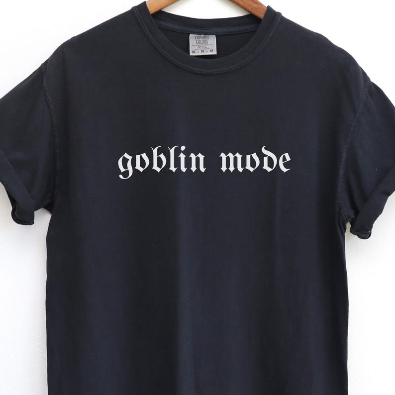 May include: Black t-shirt with the words "goblin mode" printed in white gothic font across the front. The shirt has short sleeves and a crew neck. The fabric appears to be a soft cotton blend.