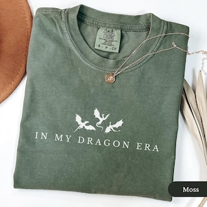 May include: A moss green Comfort Colors t-shirt with the phrase "IN MY DRAGON ERA" and three dragon silhouettes printed in white. A gold necklace with a pendant rests on the shirt.