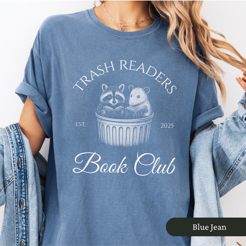 Puede incluir: Una camiseta de mezclilla azul con un gr&aacute;fico blanco de un mapache y una rata leyendo libros en un bote de basura. El texto "Trash Readers Book Club" est&aacute; impreso en la camiseta con las fechas "Est. 2025".
