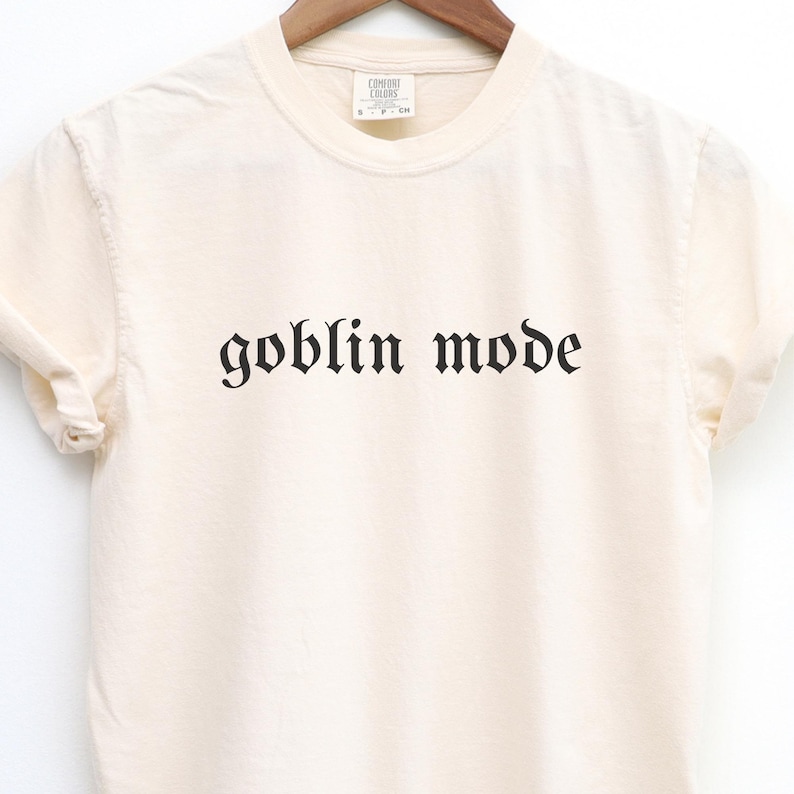 May include: Cream-colored t-shirt with the phrase "goblin mode" printed in black gothic font. The shirt is made of a soft material and has a crew neck. The shirt is hanging on a wooden hanger.