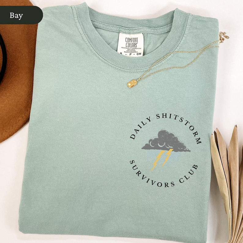 May include: A light green t-shirt with the text "DAILY SHITSTORM SURVIVORS CLUB" in a circular design. The design includes a gray cloud with yellow lightning bolts. A gold necklace with a pendant rests on the shirt.