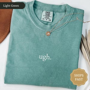May include: Light green Comfort Colors t-shirt with the word "ugh" embroidered in white. A gold-colored necklace with a pendant rests on the shirt. The shirt has a tag that reads "Comfort Colors".