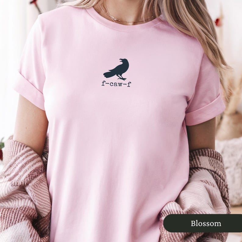 May include: A light pink t-shirt featuring a dark blue raven graphic and the text "f-caw-f". The shirt has short sleeves and a crew neck. The word "Blossom" is in a dark green rectangle at the bottom of the image.
