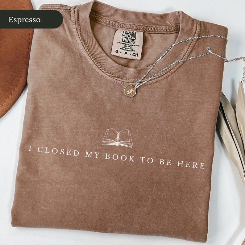 May include: Espresso-colored t-shirt with the text "I CLOSED MY BOOK TO BE HERE" and a graphic of an open book. A silver necklace with a pendant rests on the shirt. The shirt is folded and has a Comfort Colors tag.