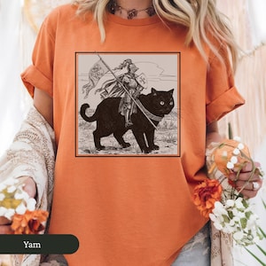 May include: Orange t-shirt with a black and white illustration of a knight riding a black cat. The illustration is framed in a rectangle and has a vintage aesthetic.