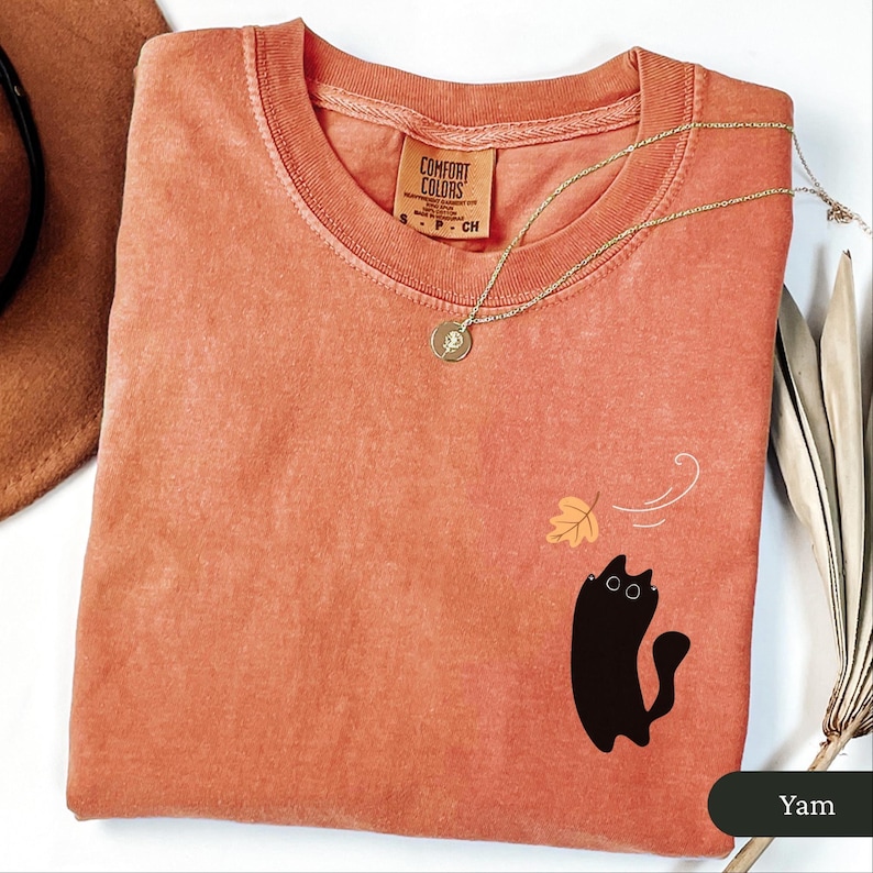 May include: A coral-coloured Comfort Colors t-shirt featuring a black cat graphic and a falling leaf design. A gold necklace with a pendant is draped on the shirt. The shirt has a tag that reads "Comfort Colors".