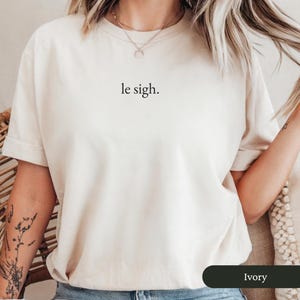Pu&ograve; includere: T-shirt color avorio con la frase "le sigh." stampata in nero. La maglietta ha maniche corte e girocollo. Il tessuto appare morbido e confortevole, adatto all'abbigliamento casual.