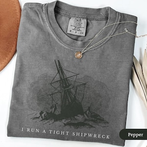May include: A gray Comfort Colors t-shirt with a graphic of a shipwreck and the text "I RUN A TIGHT SHIPWRECK". A gold-colored necklace with a pendant rests on the shirt. The shirt is folded and has a small tag.