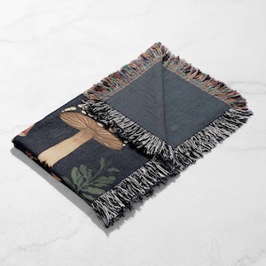 Puede incluir: Una manta plegada de color azul oscuro con un dise&ntilde;o de seta y hojas. La manta tiene un borde con flecos con una mezcla de colores. El dise&ntilde;o incluye una seta grande en tonos marrones y beige, con hojas verdes.