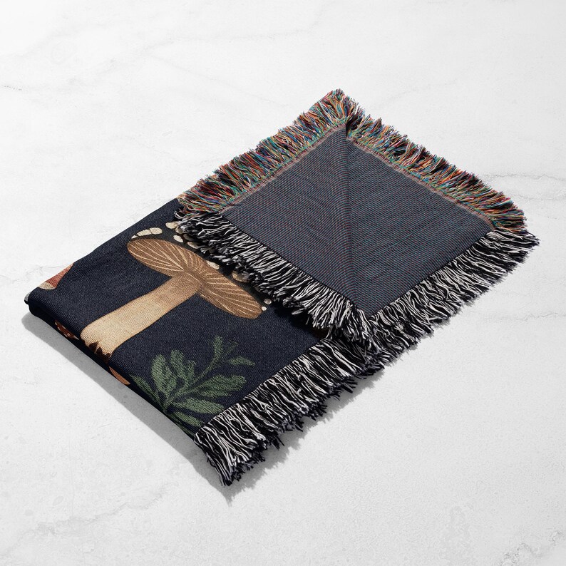 Puede incluir: Una manta plegada de color azul oscuro con un dise&ntilde;o de seta y hojas. La manta tiene un borde con flecos con una mezcla de colores. El dise&ntilde;o incluye una seta grande en tonos marrones y beige, con hojas verdes.
