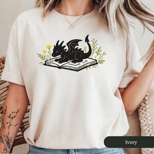 Puede incluir: Camiseta color marfil con una ilustraci&oacute;n de un drag&oacute;n negro descansando sobre un libro abierto. El drag&oacute;n est&aacute; rodeado de acentos florales amarillos y rosas. El color de la camiseta est&aacute; etiquetado como "Ivory" en la parte inferior.