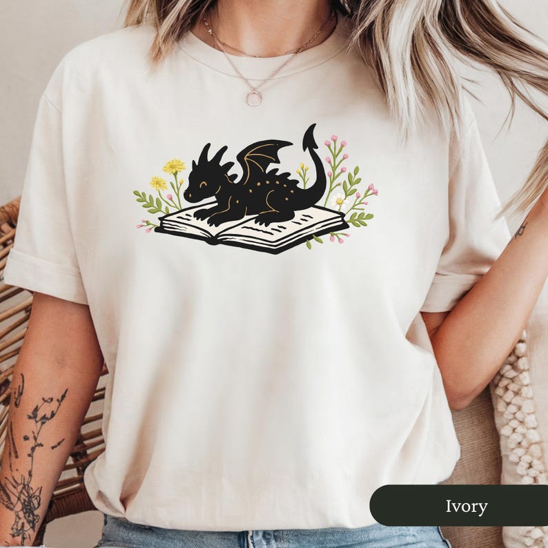 Puede incluir: Camiseta color marfil con una ilustraci&oacute;n de un drag&oacute;n negro descansando sobre un libro abierto. El drag&oacute;n est&aacute; rodeado de acentos florales amarillos y rosas. El color de la camiseta est&aacute; etiquetado como "Ivory" en la parte inferior.