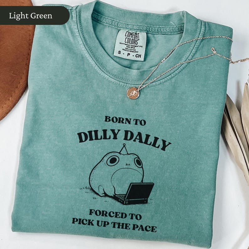 May include: Light green t-shirt with the text "BORN TO DILLY DALLY FORCED TO PICK UP THE PACE" and a cartoon frog with a laptop. The shirt has a crew neck and short sleeves. A silver necklace with a pendant is on top of the shirt.