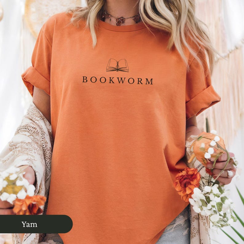 Op de afbeelding: Een oranje t-shirt met opgerolde mouwen, met een zwarte lijntekening van een open boek boven het woord "BOOKWORM". De tekst is in een eenvoudige schreefloze lettertype. Het shirt is effen van kleur en lijkt van een zacht materiaal te zijn.