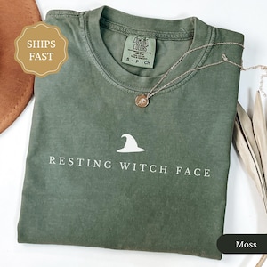 May include: A moss green t-shirt with the phrase "RESTING WITCH FACE" and a white witch hat graphic. A gold necklace with a pendant rests on the shirt. The shirt is folded and has a tag that says "Comfort Colors".
