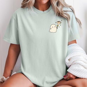 May include: A light green t-shirt featuring a cute ghost holding a magic wand, with a coffee cup and stars. The shirt is a soft, comfortable material.