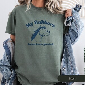 May include: Moss green t-shirt with the text "My flabbers have been gasted" and a blue horse illustration. The shirt is a crew neck style and has short sleeves. The shirt is made of a soft material.