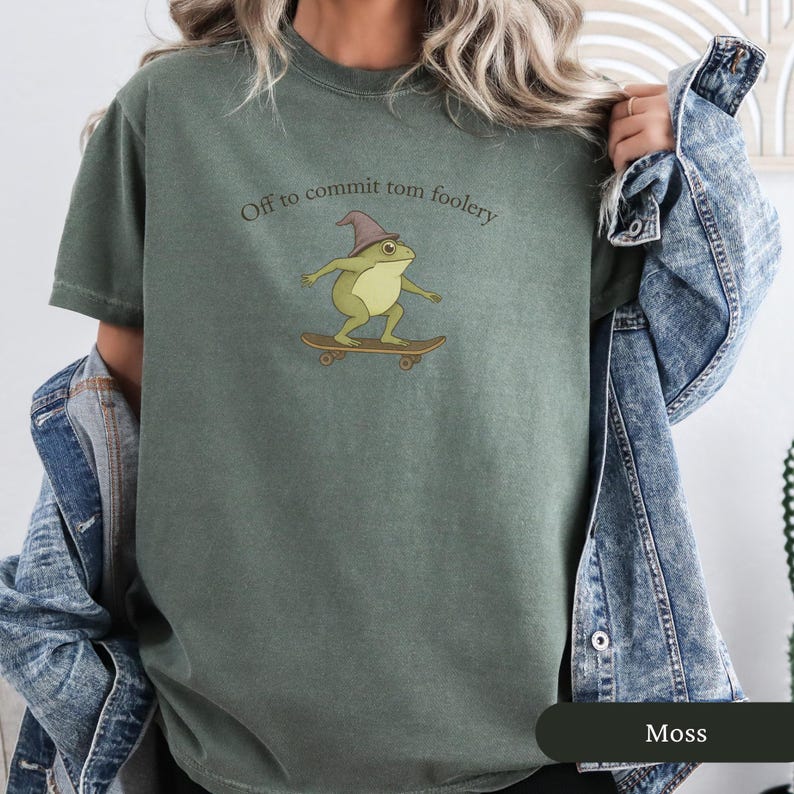 Puede incluir: Camiseta verde musgo con un dibujo de una rana de dibujos animados en monopat&iacute;n, con un sombrero de mago. El texto "Off to commit tom foolery" est&aacute; sobre la rana. La camiseta es de corte holgado.