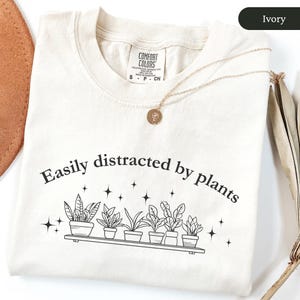 May include: Cream-coloured t-shirt featuring the phrase "Easily distracted by plants" and a graphic of potted plants. A gold necklace with a circular pendant is on the shirt. The t-shirt is made of a soft material.