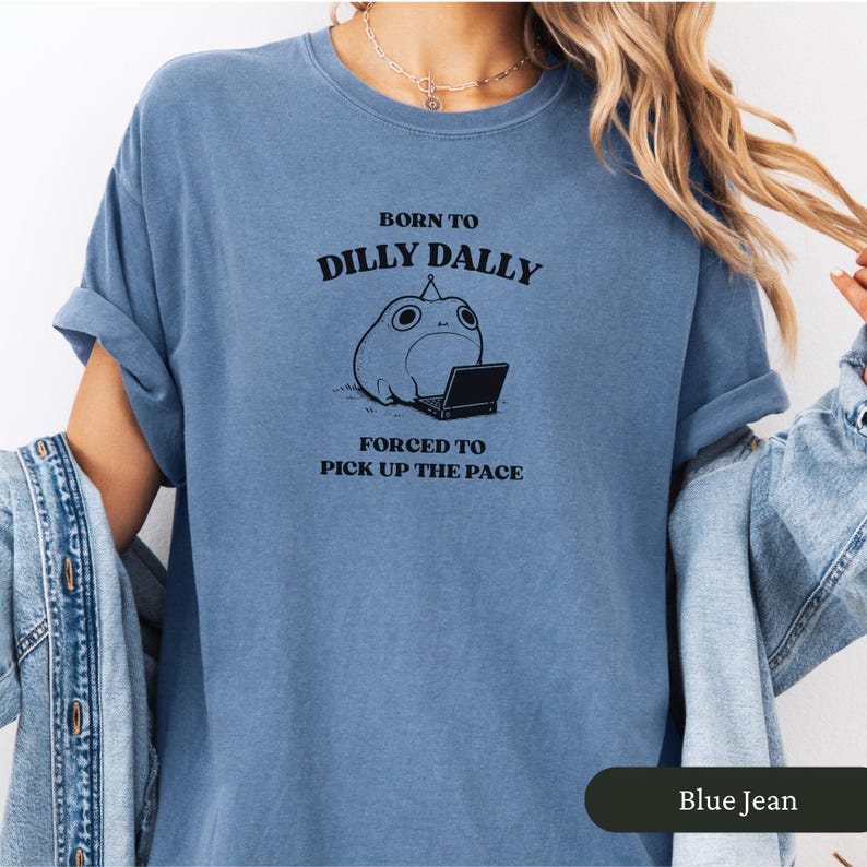 May include: Blue jean-colored t-shirt with the text "BORN TO DILLY DALLY FORCED TO PICK UP THE PACE" and a cartoon frog with a laptop. The shirt has rolled-up sleeves and a relaxed fit.