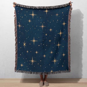 Puede incluir: Una manta azul marino con un patr&oacute;n repetido de estrellas doradas. La manta tiene un borde con flecos en tonos marrones y beige. Las estrellas var&iacute;an en tama&ntilde;o y est&aacute;n espaciadas uniformemente sobre la superficie de la manta.