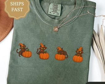 May include: A moss green t-shirt featuring four cartoon black cats wearing witch hats, each perched on an orange pumpkin. The shirt has a crew neck and short sleeves. A gold necklace is draped across the shirt.
