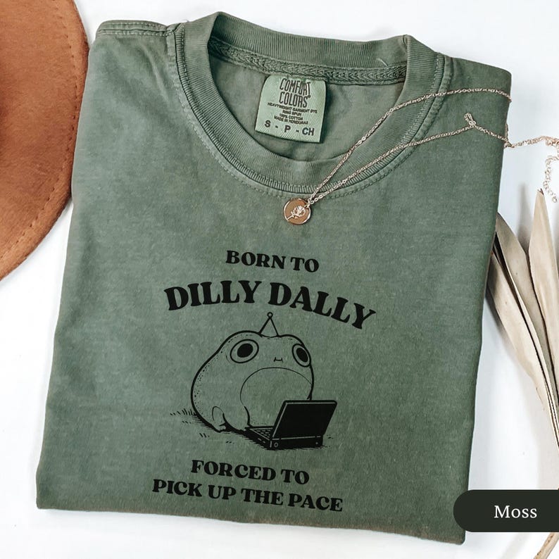 May include: Moss green t-shirt with the text "BORN TO DILLY DALLY FORCED TO PICK UP THE PACE" and a cartoon frog with a laptop. A gold necklace with a pendant is on the shirt.