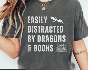Puede incluir: Camiseta gris oscuro con el texto blanco "EASILY DISTRACTED BY DRAGONS & BOOKS". Un diseño de dragón y libro adorna la prenda. Esta camiseta informal tiene un estilo cómodo.