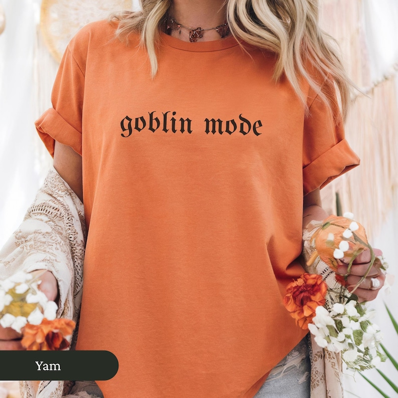 May include: Orange t-shirt with rolled-up sleeves featuring the phrase "goblin mode" in a gothic font. The shirt is a solid color and appears to be made of a soft material. The overall theme is casual wear.