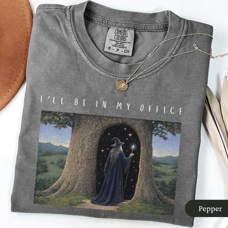 Op de afbeelding: Pepperkleurig t-shirt met de tekst "I'll Be In My Office". De afbeelding toont een tovenaar in een blauwe mantel en hoed in een boomdeur. Een zilveren ketting met een kleine hanger ligt op het shirt. Het shirt heeft een Comfort Colors label.